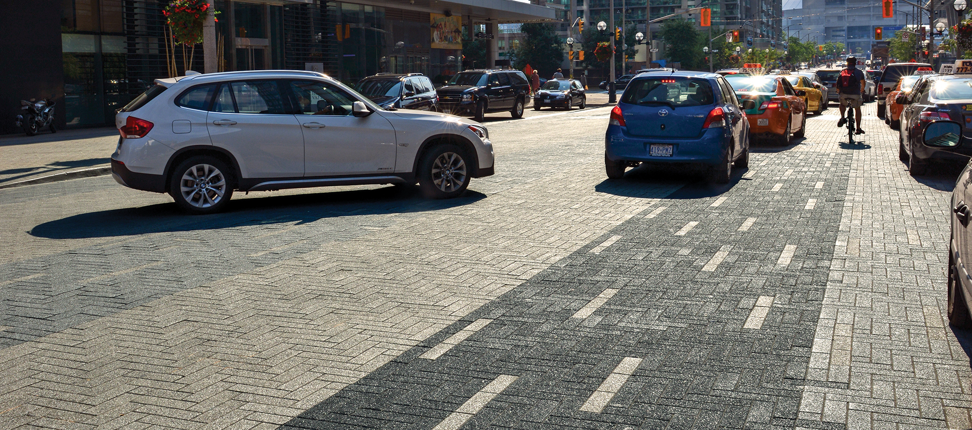 Cars and cyclists share a busy Toronto street paved with Promenade plank pavers using different colors to delineate lanes.
