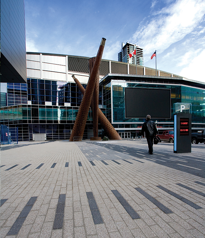A business person walks past an abstract statue in front of Scotia Bank Arena on a plaza of Series pavers with line patterns.