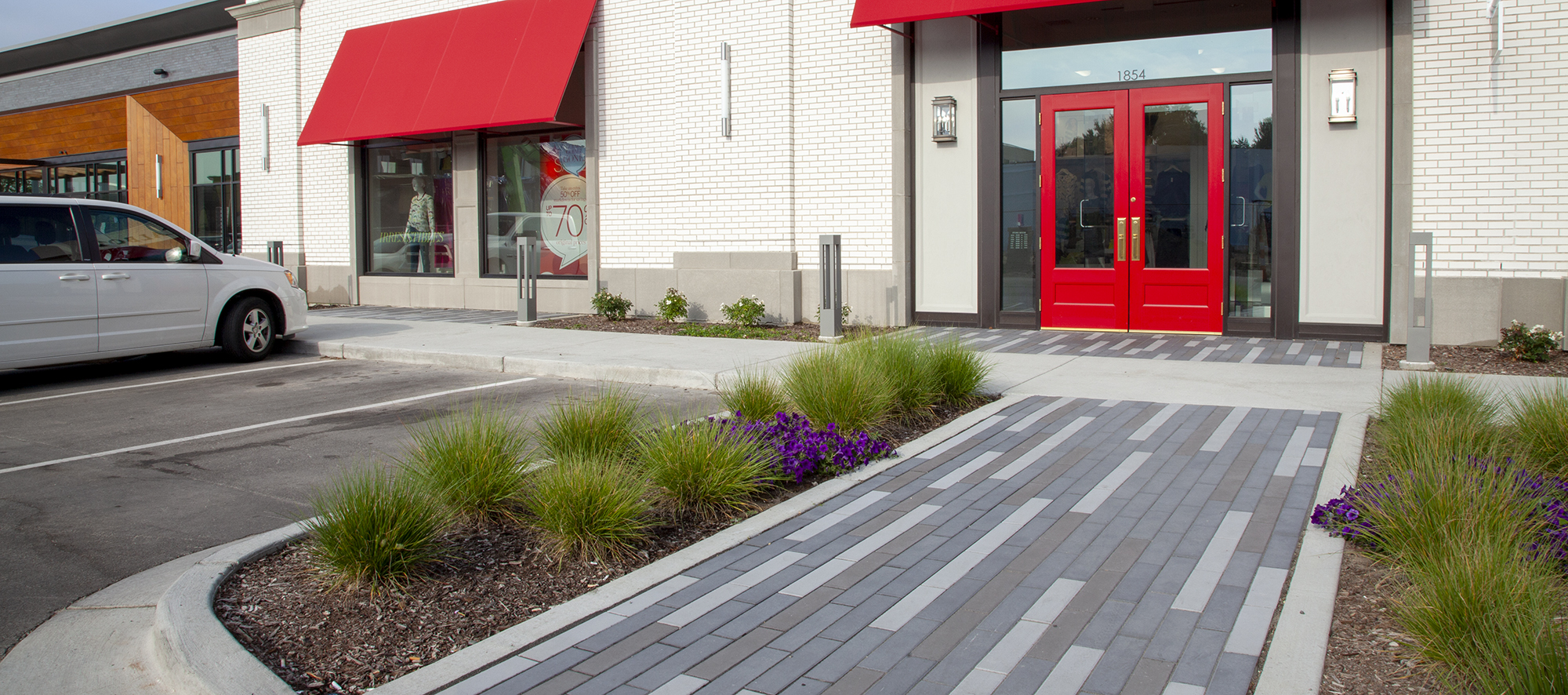 Beside a parking lot a path paved with tri-color Promenade Plank Pavers, bordered by gardens, leads to the red doors of a shopping center.