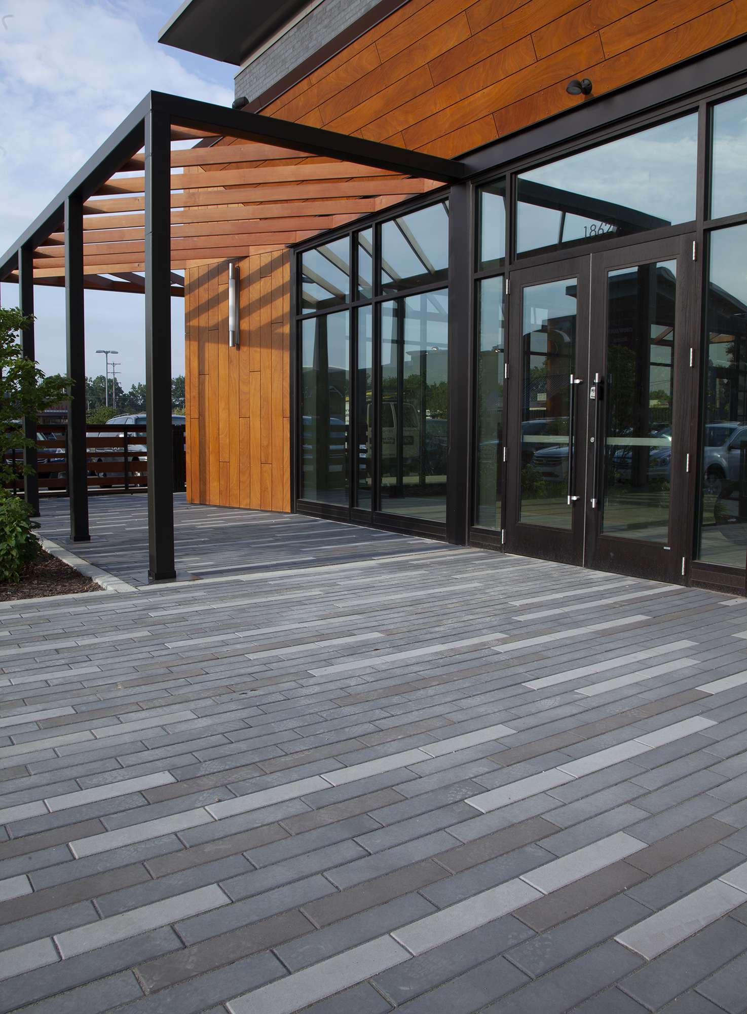 The entry to a wood panel and glass building with a modern pergola is paved with Promenade Plank Pavers in three colors in linear patterns.