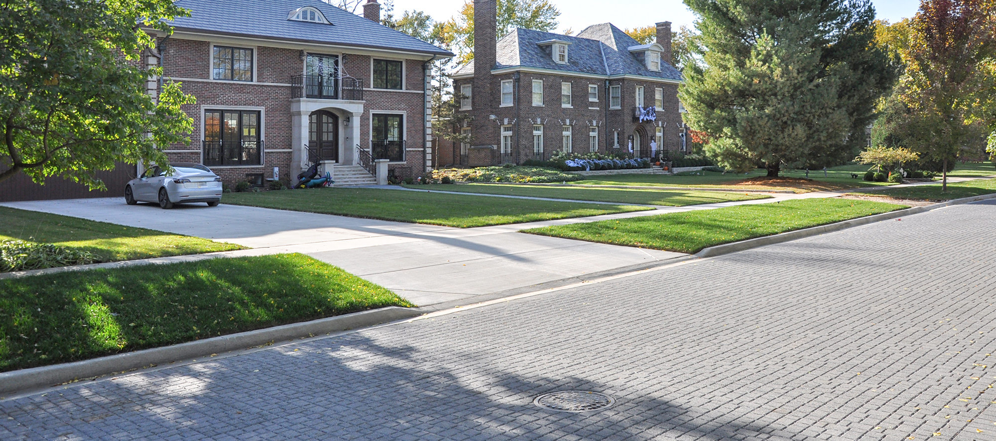 A serene streetscape showcases a stunning light grey Unilock paver roadway that leads to the driveways of two exceptional homes.