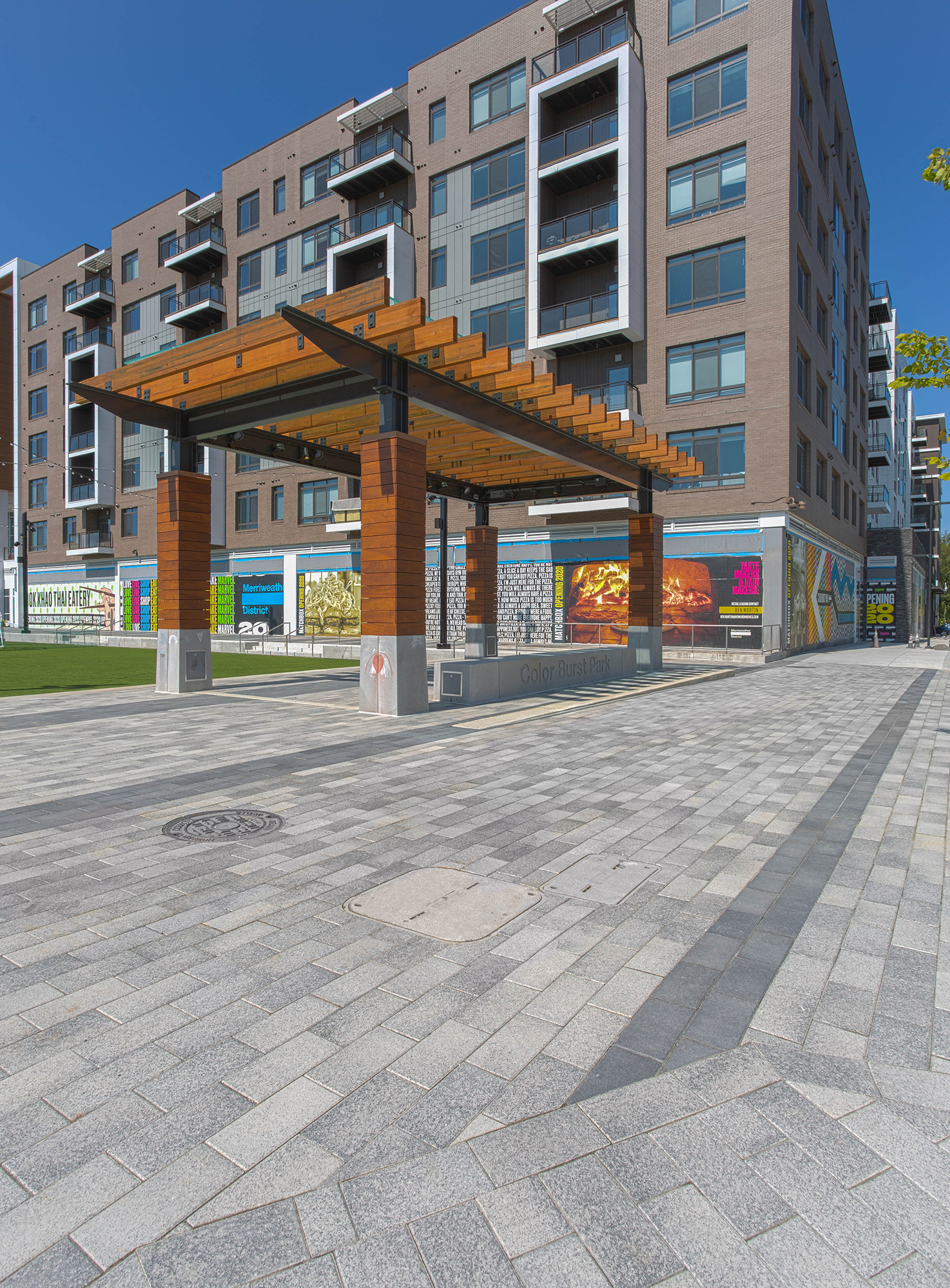 Two-toned Rectangle pavers lead to a wooden pergola for shade, followed by a colorful advertising display along the building's exterior.